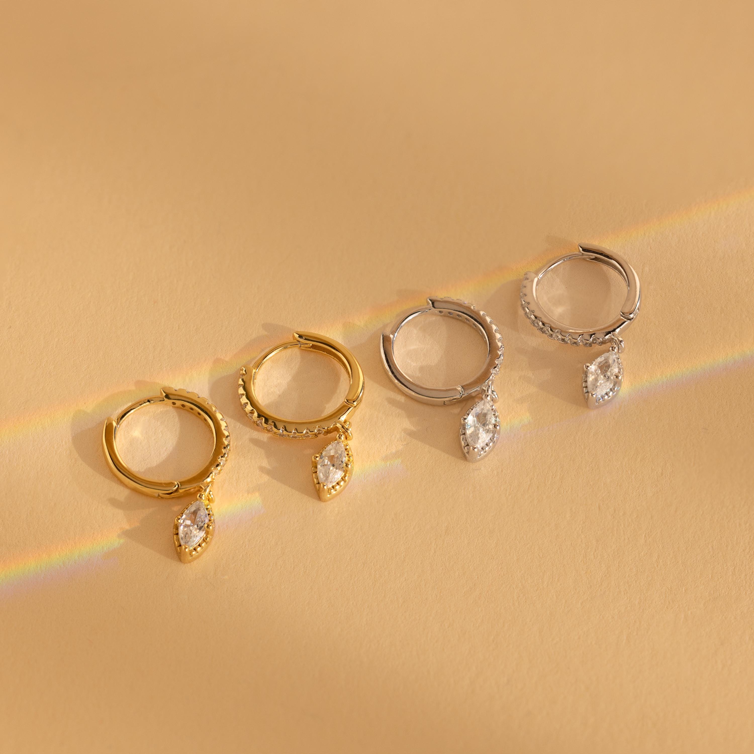 Two pairs of elegant earrings—one gold, one silver—feature a dangling marquise-cut gem, inspired by the Marquise Pave Drop Huggies, and are displayed on a beige surface.