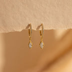 The Marquise Pave Drop Huggies are elegant gold hoop earrings with sparkling crystal drops, beautifully displayed on textured beige paper against a soft background.
