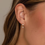 Close-up of an ear adorned with Butterfly Drop Huggies, gold earrings featuring a floral motif and two small dangling gems, reminiscent of diamond accent earrings.