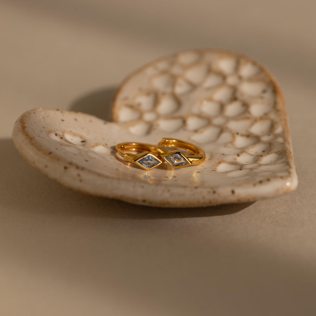 Two gold rings with square gemstones sit on a textured heart-shaped ceramic dish, evoking the refined style of Rhombus Birthstone Huggies.