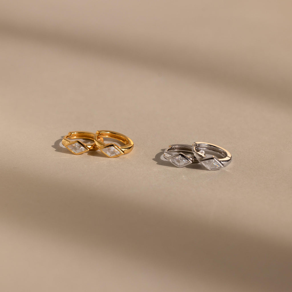 Two pairs of huggie earrings on a neutral background: one in gold and the other in silver, each featuring a clear rhombus-cut diamond-like birthstone, softly illuminated by natural light