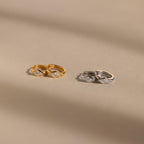 Two pairs of huggie earrings on a neutral background: one in gold and the other in silver, each featuring a clear rhombus-cut diamond-like birthstone, softly illuminated by natural light