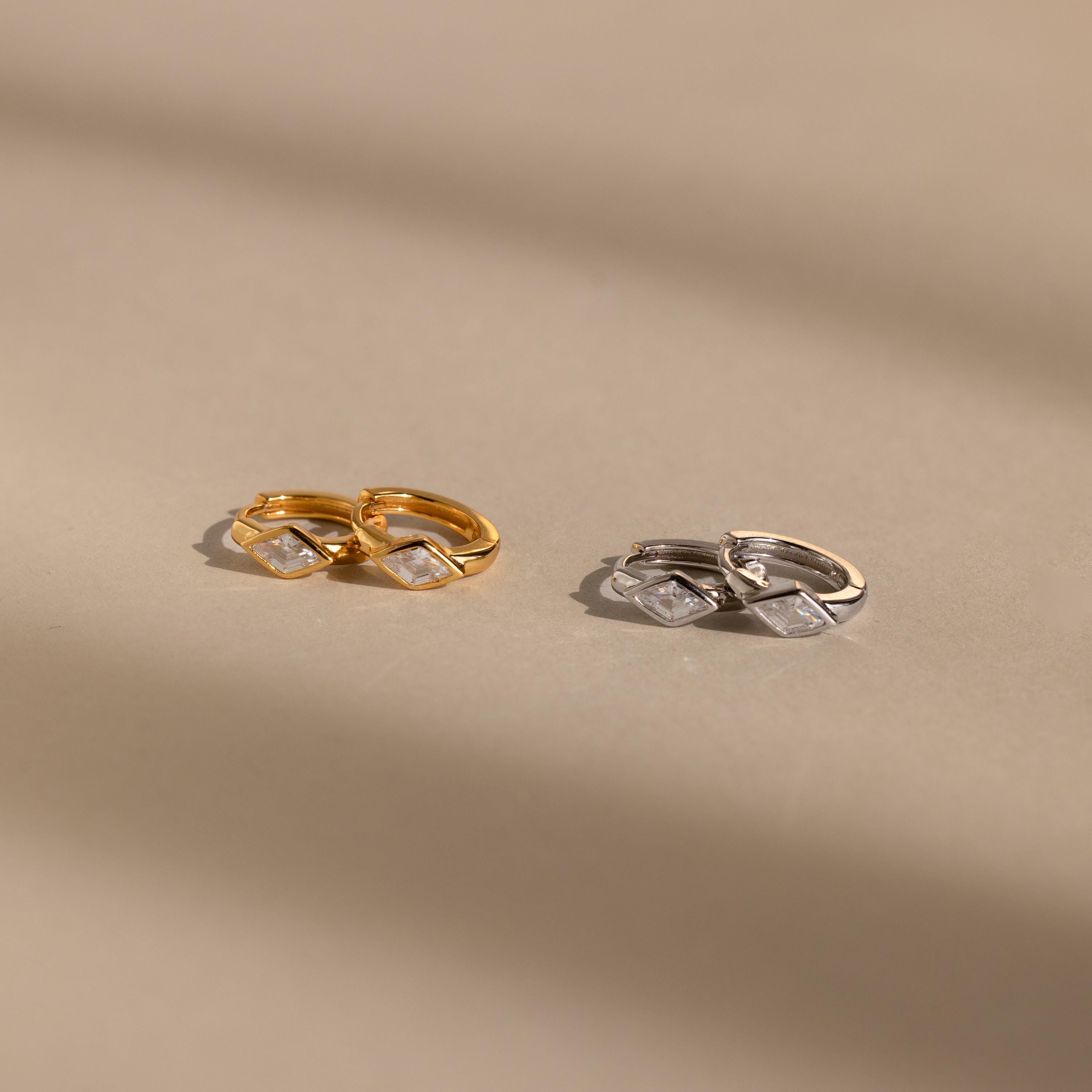 Two pairs of huggie earrings on a neutral background: one in gold and the other in silver, each featuring a clear rhombus-cut diamond-like birthstone, softly illuminated by natural light