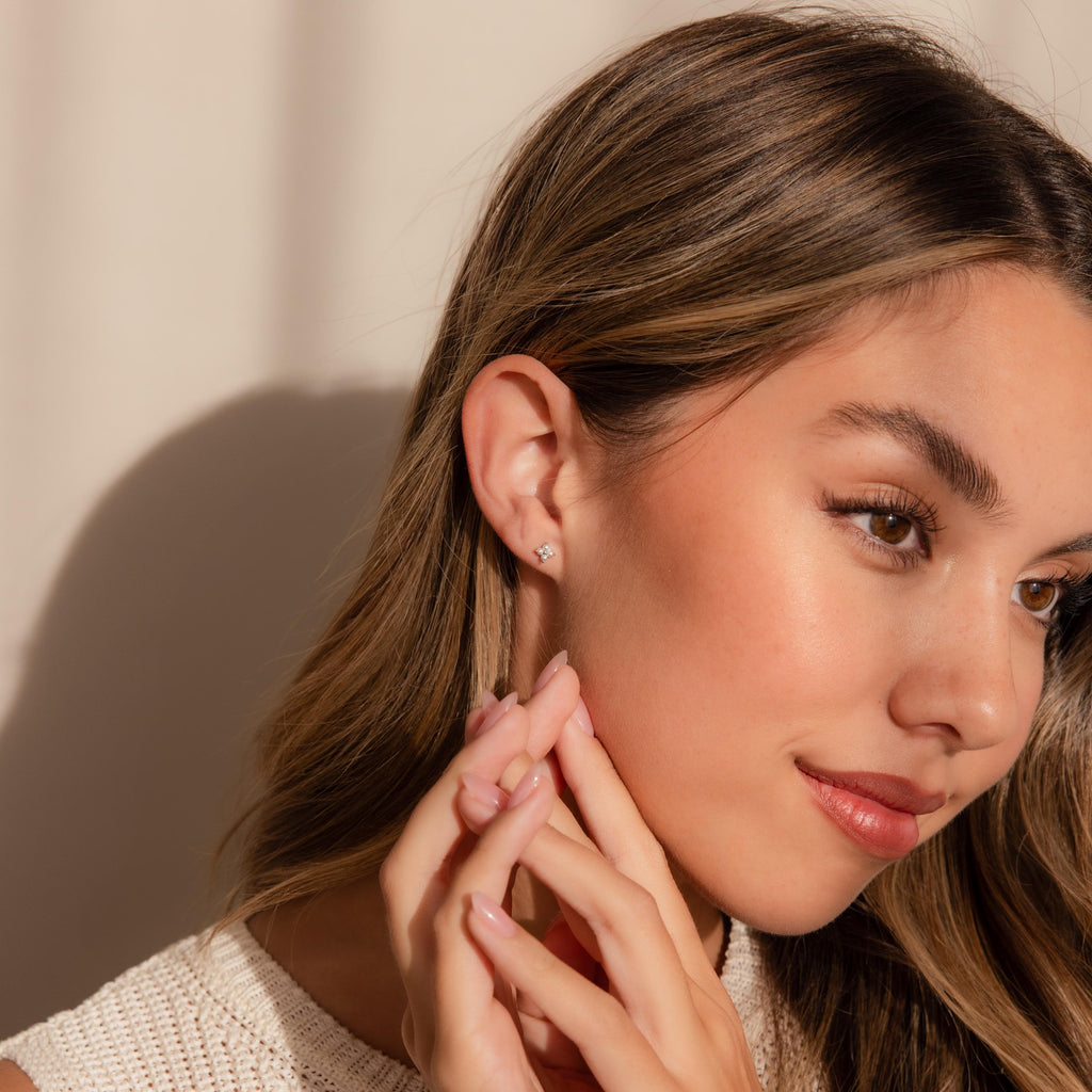 A woman with long brown hair wears Pearl Ivy Flower Studs, gently touching her face while gazing to the side.