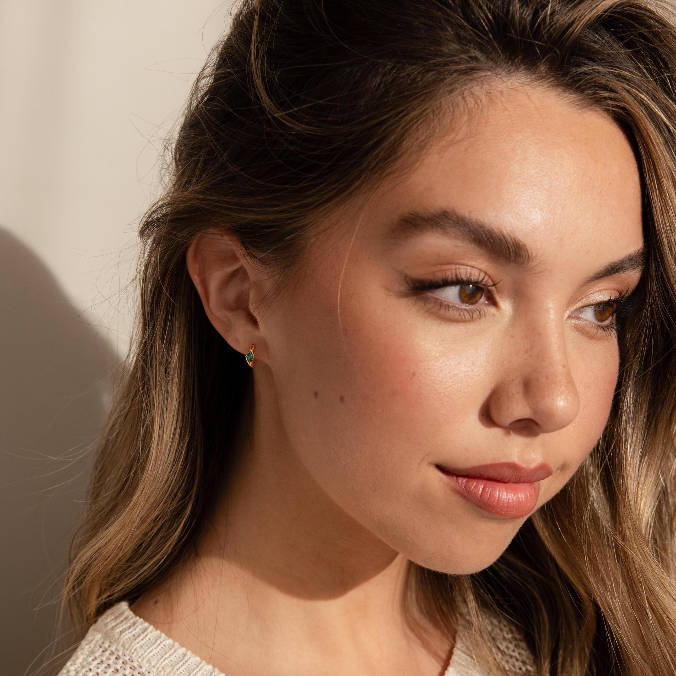 A woman facing forward, wearing a small gold huggie earring with a green rhombus emerald stone