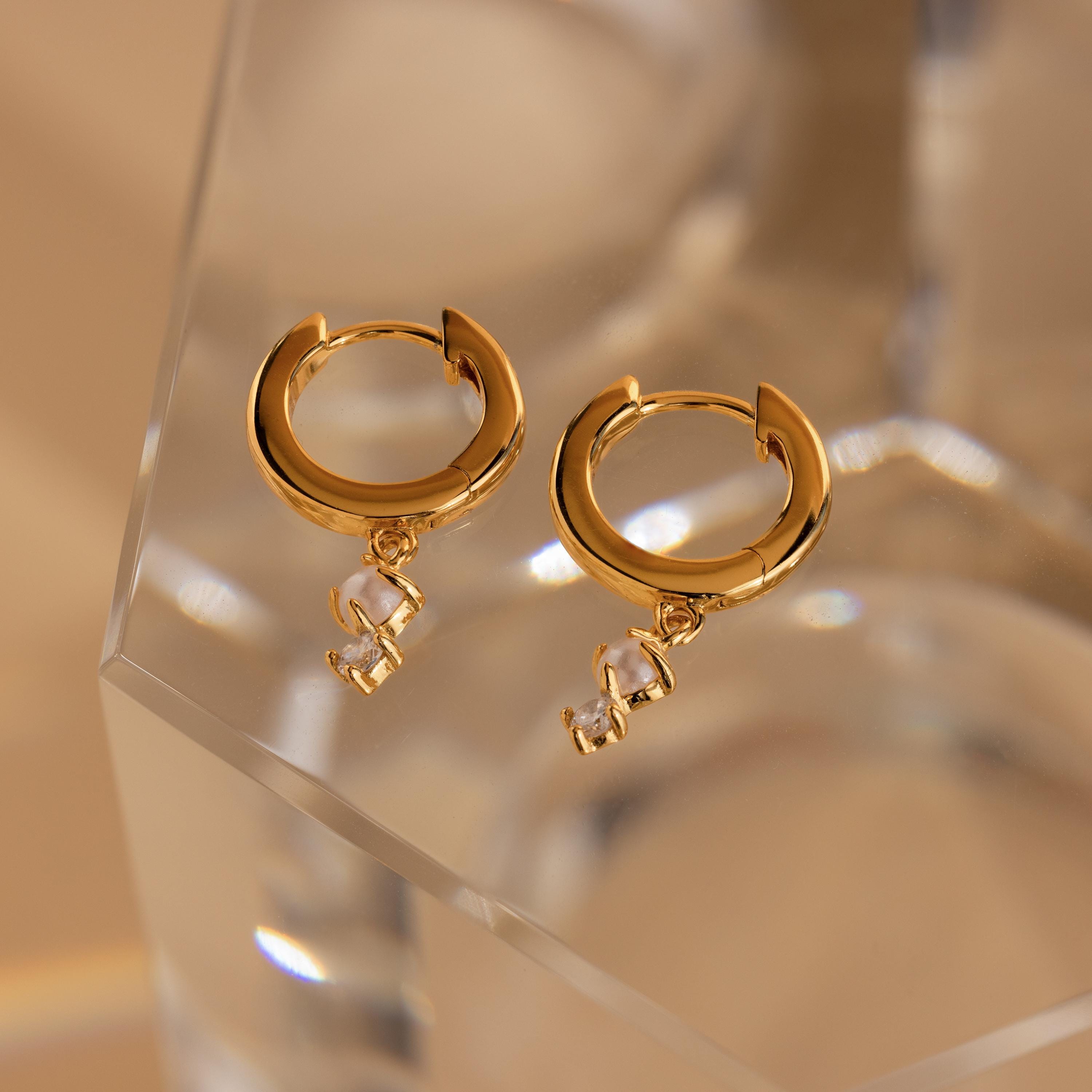 Dangling Pearl Diamond Huggies: Elegant gold hoop earrings with pearl and diamond accents, featuring dangling letter charms, add a charming twist to classic styles. Displayed on a clear faceted surface for extra sparkle.