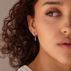 Woman with curly hair wears small silver hoop earrings featuring a dangling charm, gazing to the side. The style captures the timeless elegance of the Dangling Pearl Diamond Huggies.