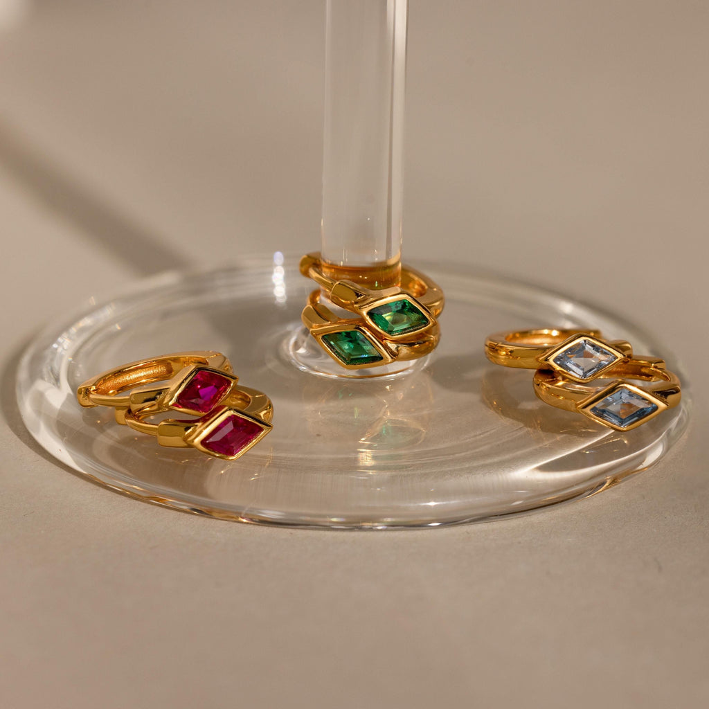 Three sets of gold huggie earrings with rhombus-shaped birthstones displayed on a clear glass stand—emerald green, ruby red, and aquamarine blue stones—bathed in warm light.