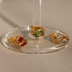 Three sets of gold huggie earrings with rhombus-shaped birthstones displayed on a clear glass stand—emerald green, ruby red, and aquamarine blue stones—bathed in warm light.