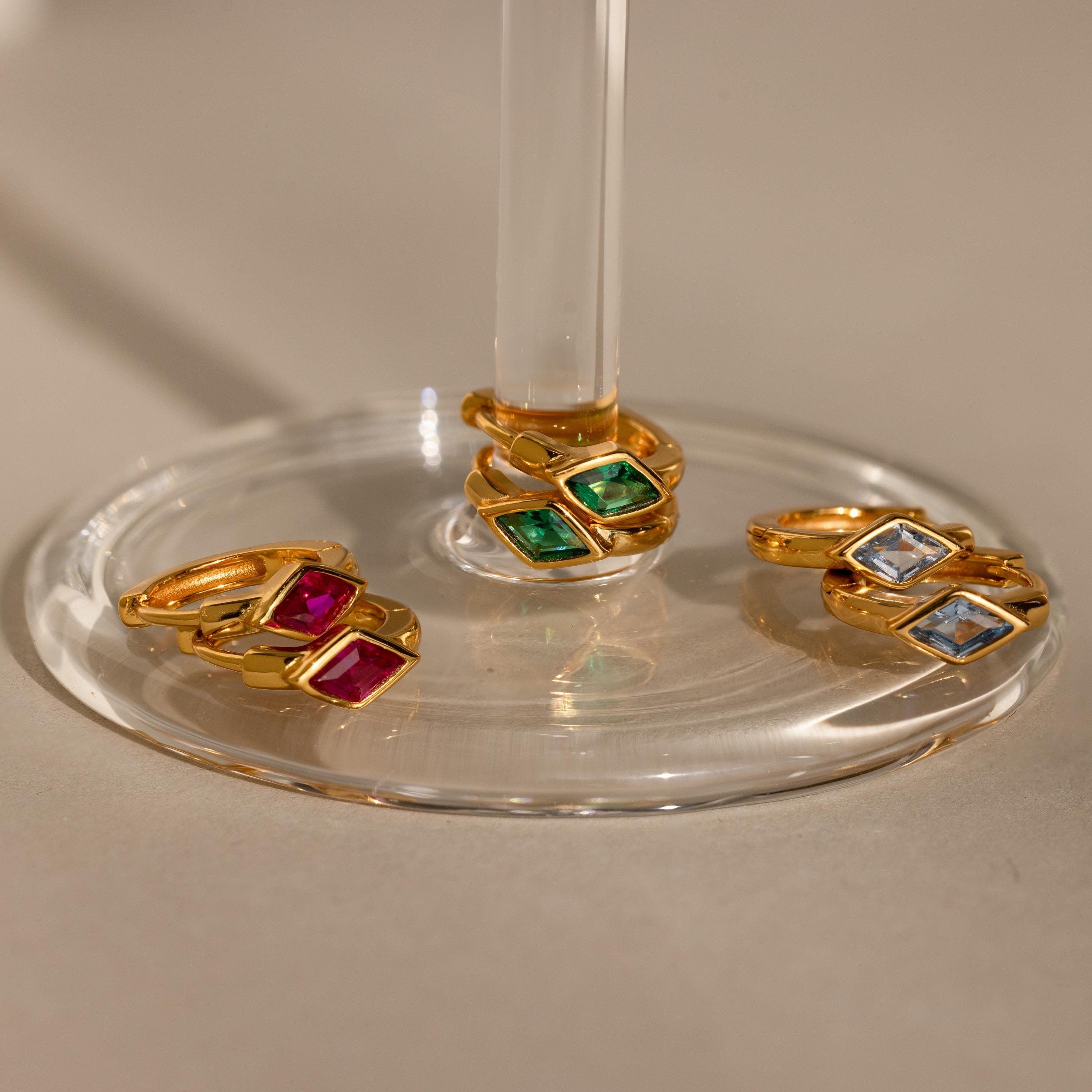 Three sets of gold huggie earrings with rhombus-shaped birthstones displayed on a clear glass stand—emerald green, ruby red, and aquamarine blue stones—bathed in warm light.