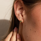 A close-up of a woman’s ear wearing a single gold huggie earring featuring a vivid green rhombus-shaped emerald birthstone, with sunlight softly highlighting the contours of her ear and hand.