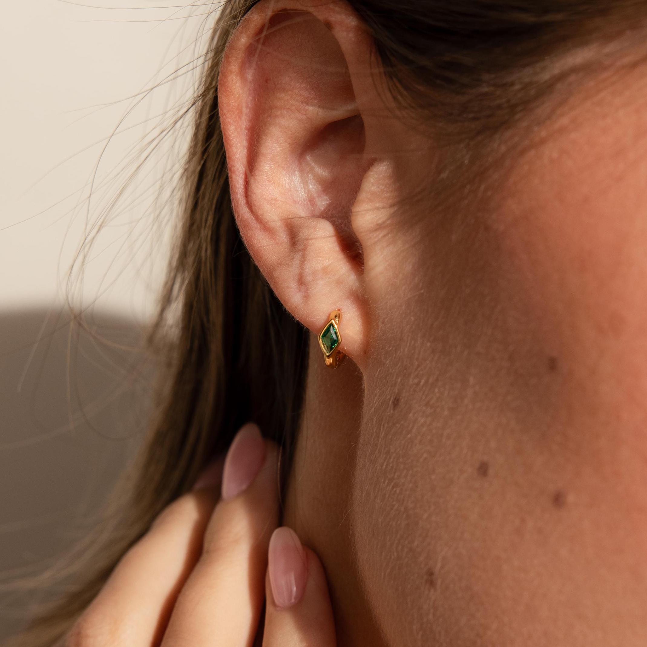 A close-up of a woman’s ear wearing a single gold huggie earring featuring a vivid green rhombus-shaped emerald birthstone, with sunlight softly highlighting the contours of her ear and hand.