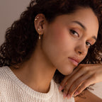 A woman with curly hair wears Dangling Pearl Diamond Huggies earrings, a nose ring, and a white knit top as she gazes thoughtfully.
