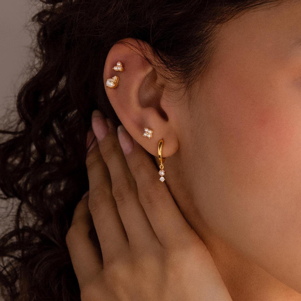 A close-up of a woman's ear adorned with Dangling Pearl Diamond Huggies in 18K Gold and multiple gold and diamond earrings, her hand gently touching her neck.
