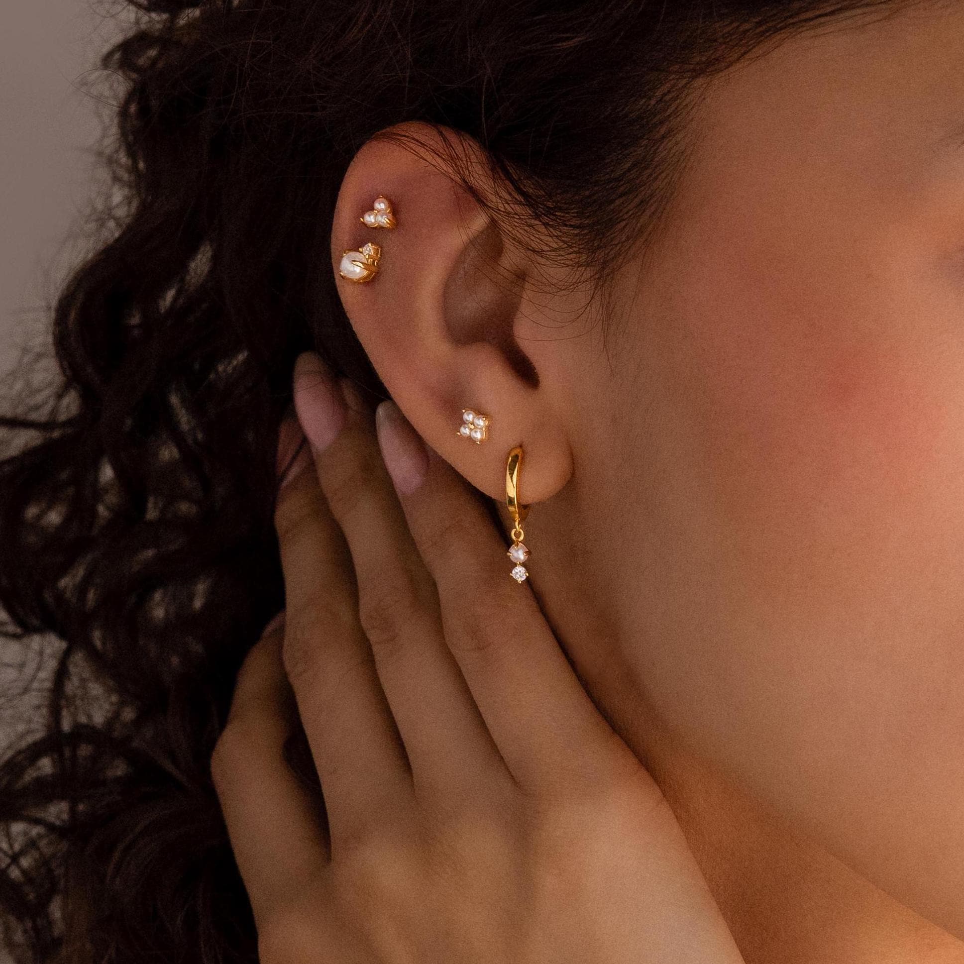 A close-up of a woman's ear adorned with Dangling Pearl Diamond Huggies in 18K Gold and multiple gold and diamond earrings, her hand gently touching her neck.