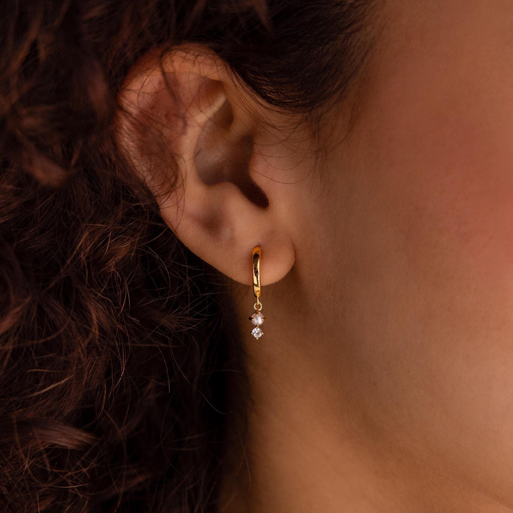 Close-up of an ear with Dangling Pearl Diamond Huggies in 18K Gold, featuring elegant hoops with two small dangling diamonds, against curly brown hair.