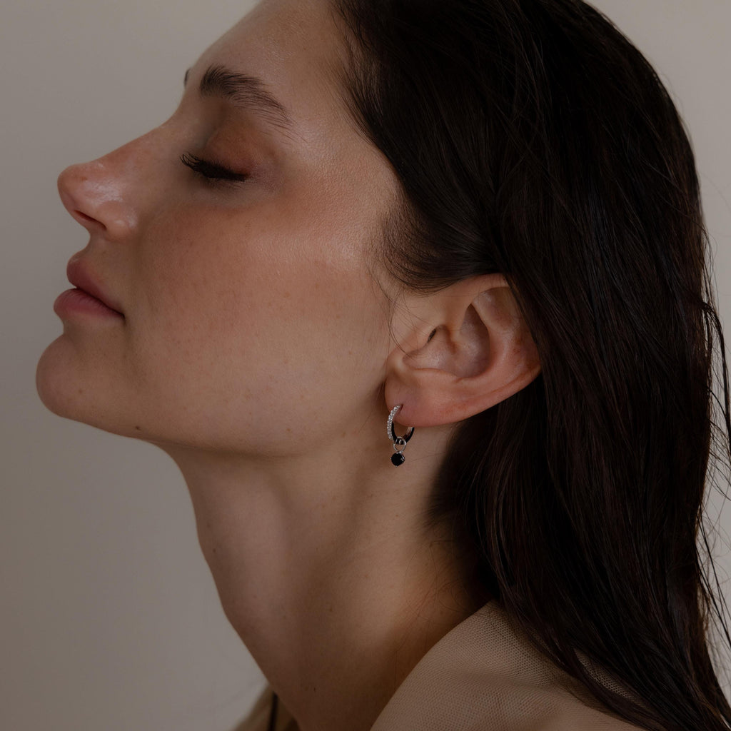 Woman in profile with closed eyes wears Black Diamond Drop Huggies—elegant hoop earrings featuring a small black bead—set against a neutral background.