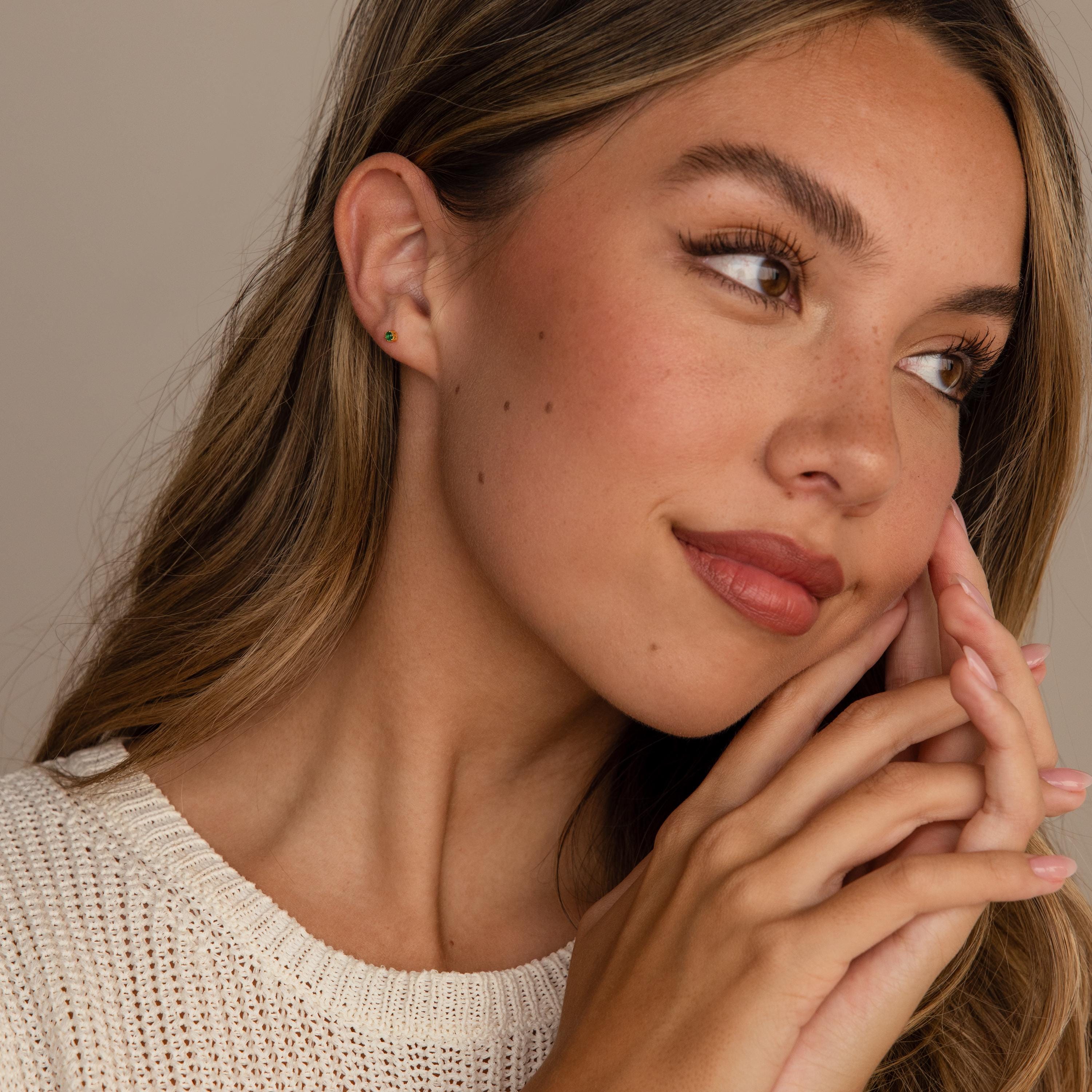 Young woman with long hair and subtle makeup, wearing a cream knit top and elegant Emerald Flatback Studs, looking off to the side smiling.