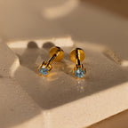 Aquamarine Flatback Studs with blue gemstones are displayed on a cream-colored textured surface under warm lighting.