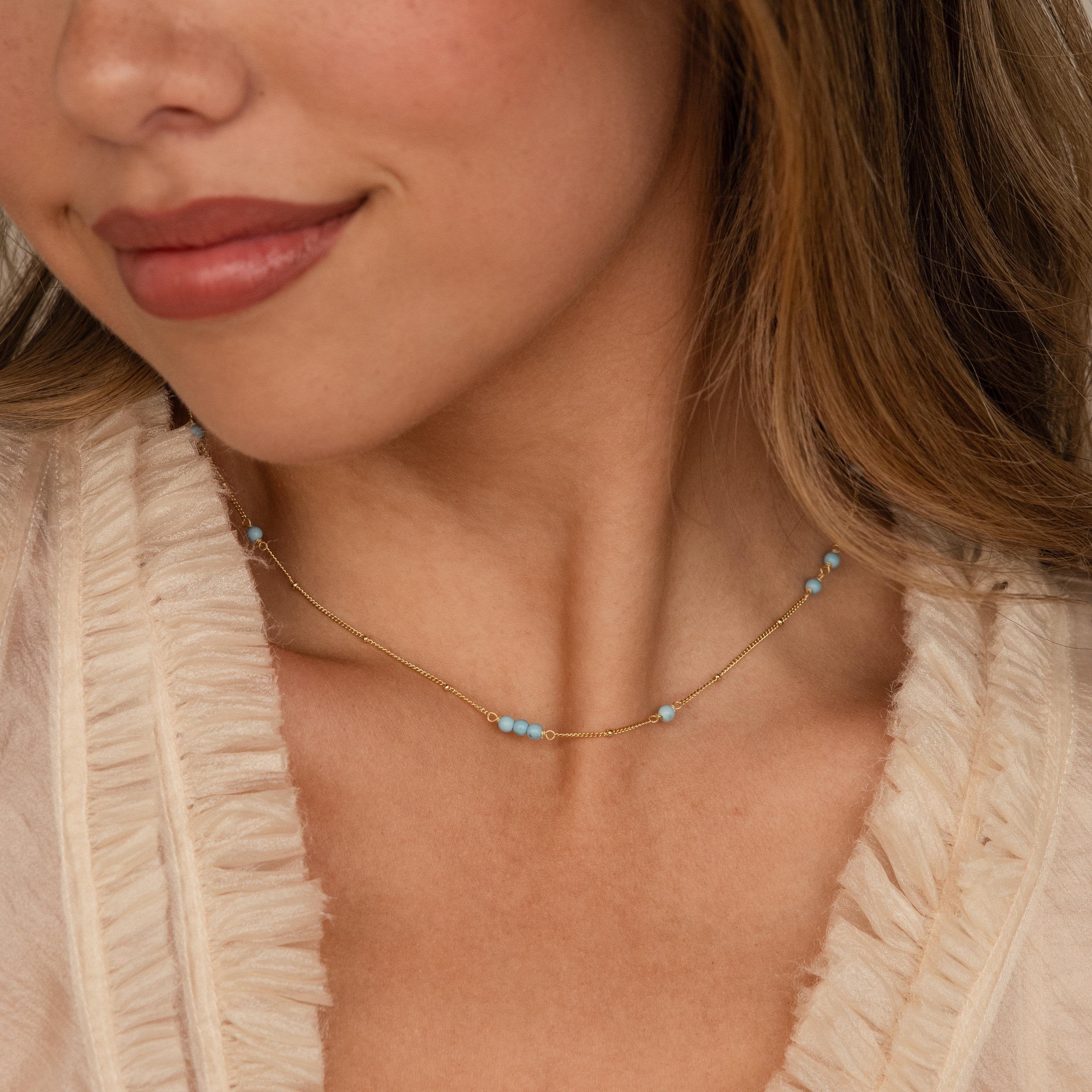 A woman wears the Turquoise Satellite Necklace, featuring small turquoise beads, elegantly styled over a cream ruffled top.