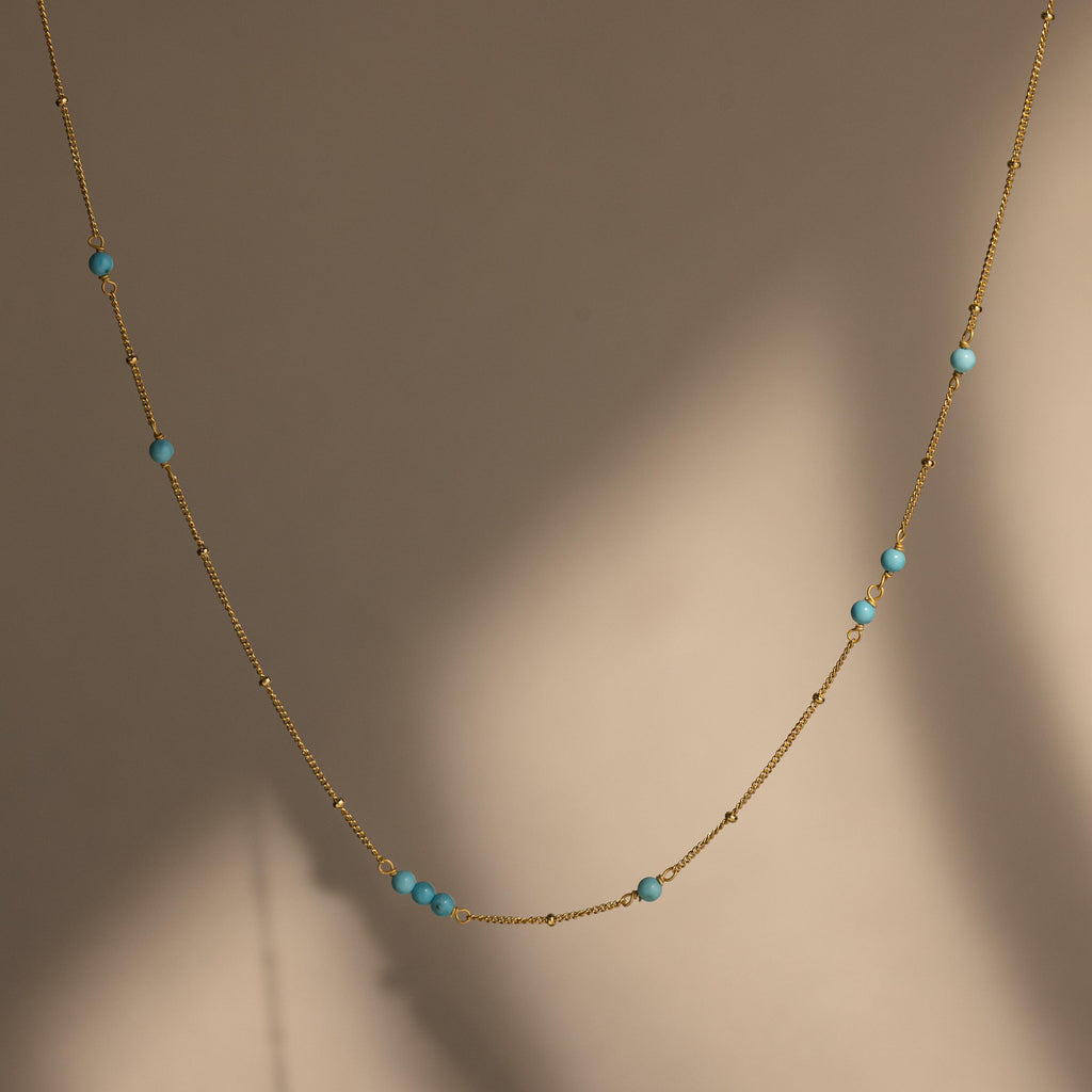 The Turquoise Satellite Necklace features delicate gold layers and small turquoise beads, shining beautifully against a softly lit neutral background.