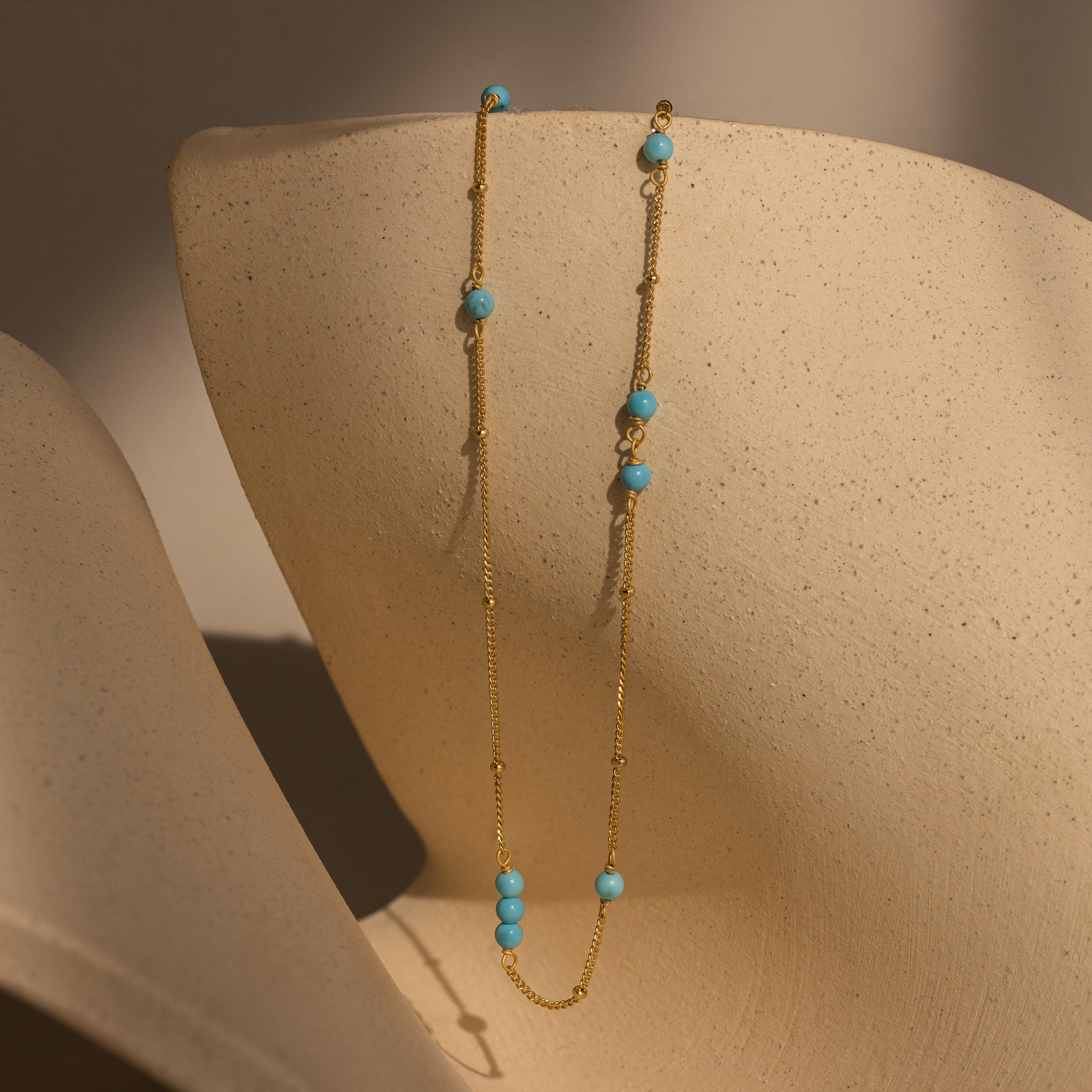 The Turquoise Satellite Necklace features delicate turquoise beads and looks stunning on a beige textured bust with soft shadows—an ideal layering piece for any jewelry collection.