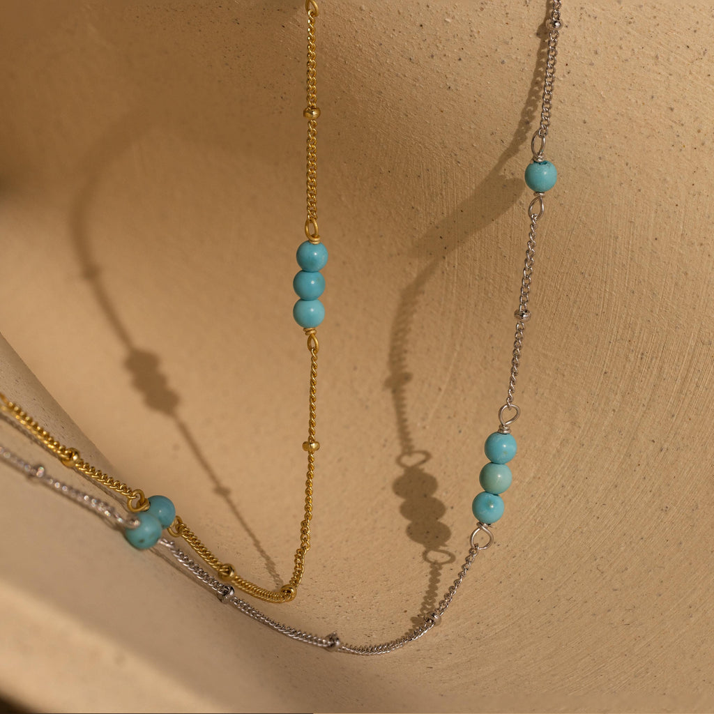 The Turquoise Satellite Necklace, featuring gold and silver layers with turquoise beads, is displayed on a textured beige surface.