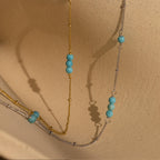 The Turquoise Satellite Necklace, featuring gold and silver layers with turquoise beads, is displayed on a textured beige surface.