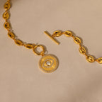 Evil Eye Toggle Necklace featuring a gold mariner chain with a circular sunburst pendant and toggle clasp, shown on a beige background.