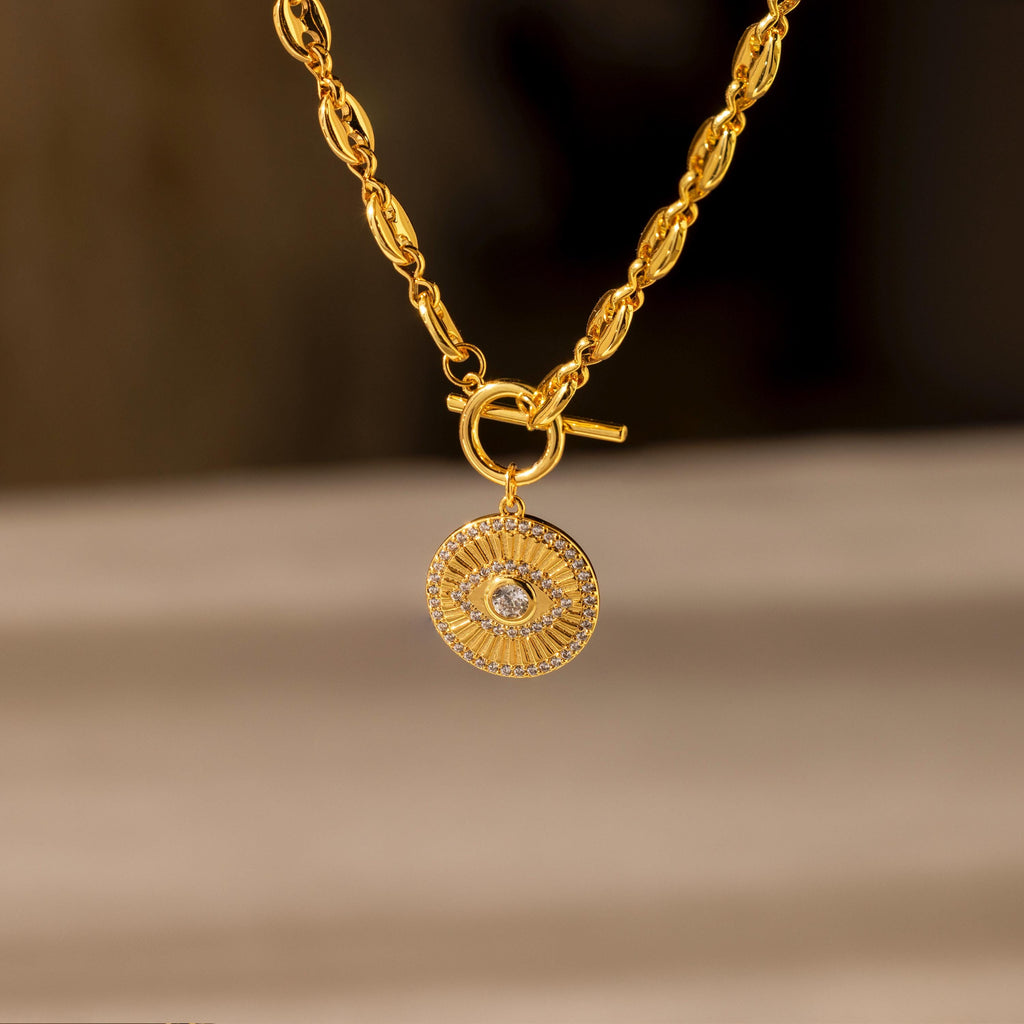 The Evil Eye Toggle Necklace features a gold mariner chain with a round, textured pendant and central clear gemstone, evoking an elegant diamond evil eye charm.