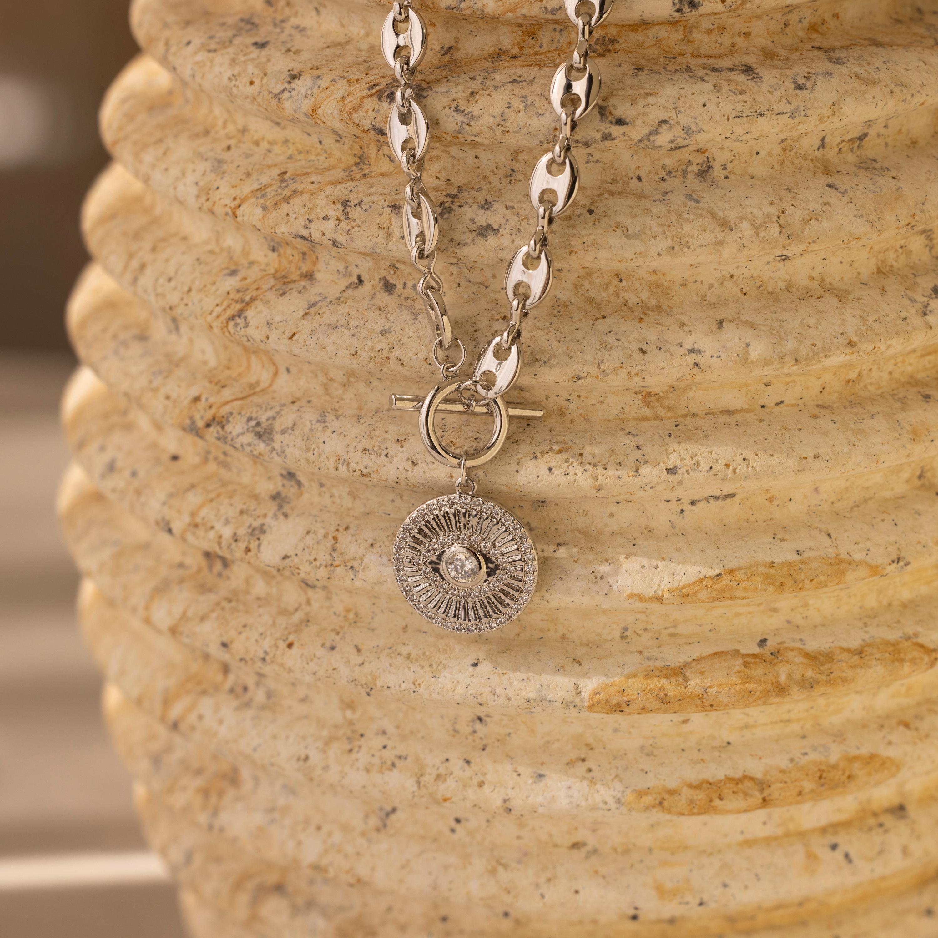 Evil Eye Toggle Necklace featuring a silver mariner chain and round, textured pendant, displayed on a textured beige stone surface.