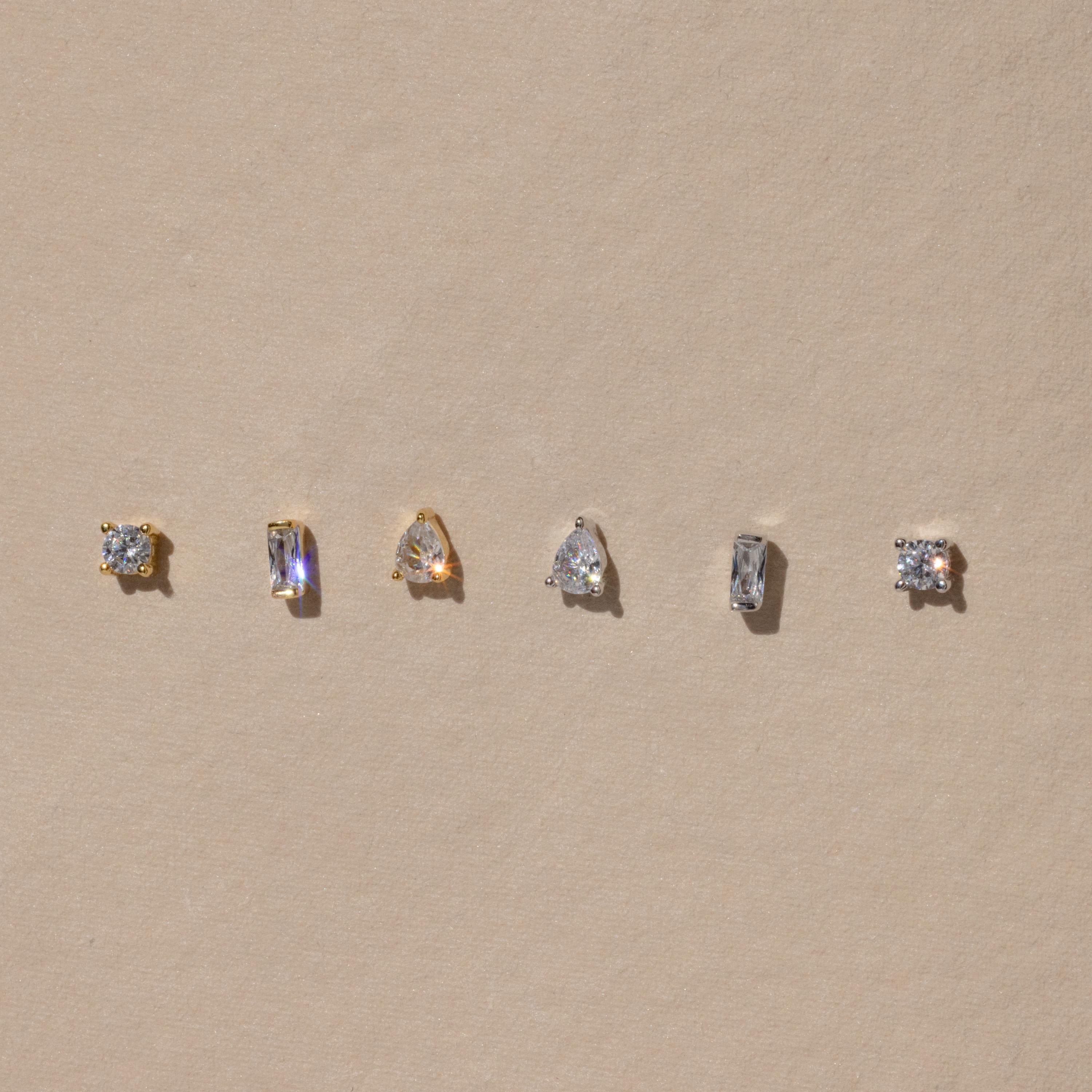 The Mixed Diamond Flat Back Stud Set features five small gold and silver stud earrings, including flat back and mixed diamond designs, displayed in a row on a beige background.