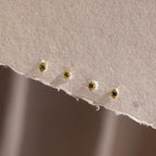 Four gold geometric flat back studs in emerald and sapphire arranged along the edge of a soft, textured paper background.