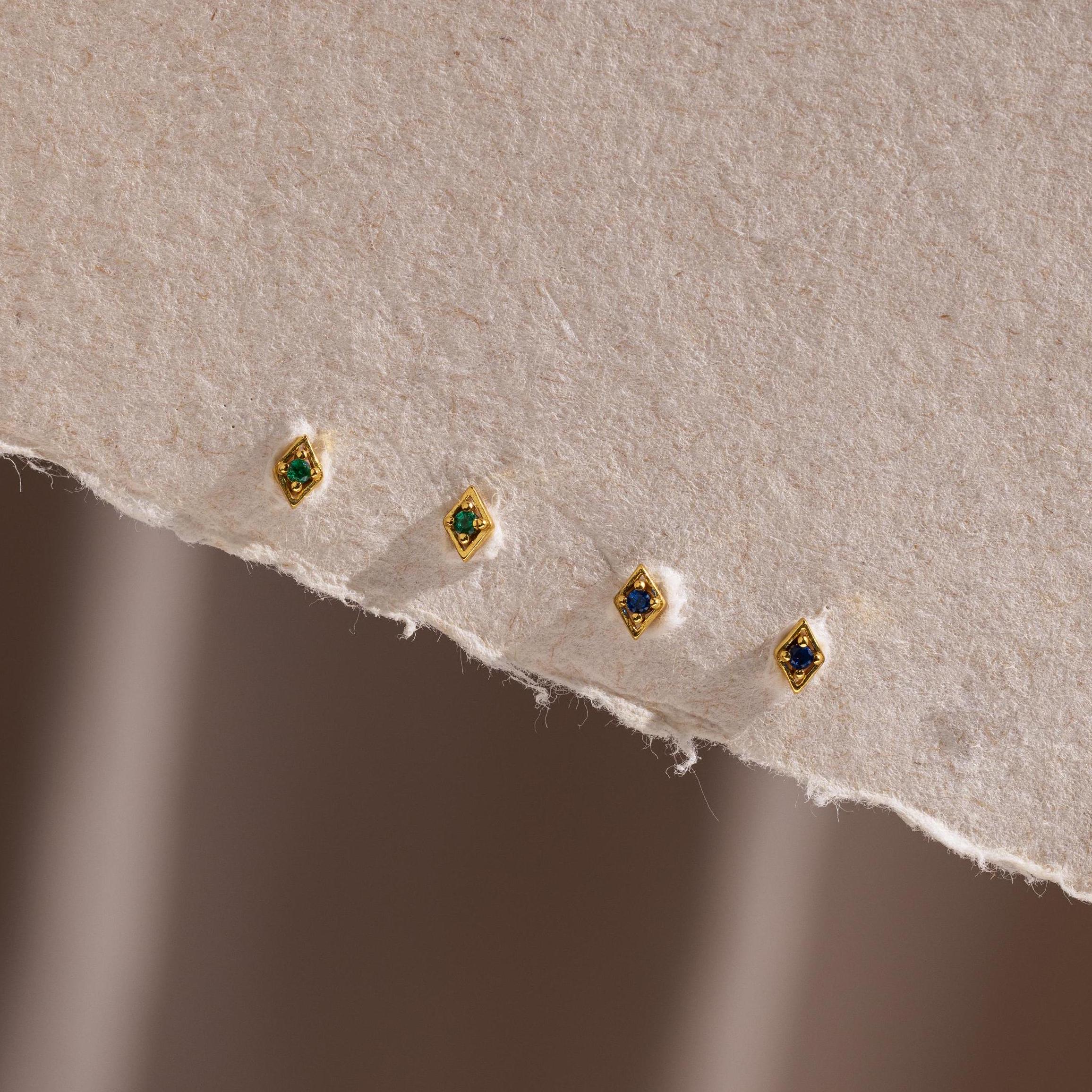 Four gold geometric flat back studs in emerald and sapphire arranged along the edge of a soft, textured paper background.