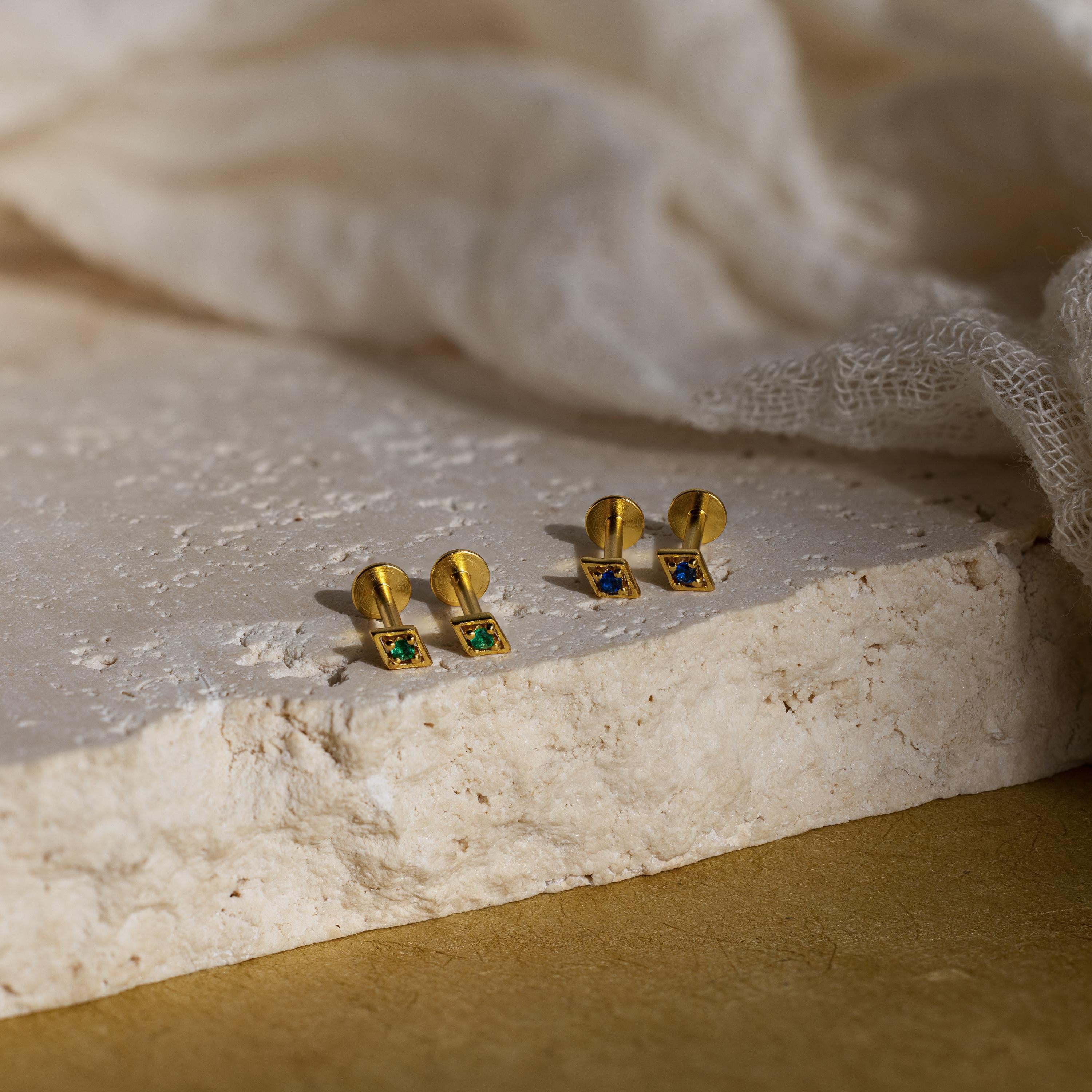 Two pairs of Geometric Birthstone Flat Back Studs, with square green and blue stones, rest on a textured stone surface—customizable earrings perfect for adding a personal touch to your style.