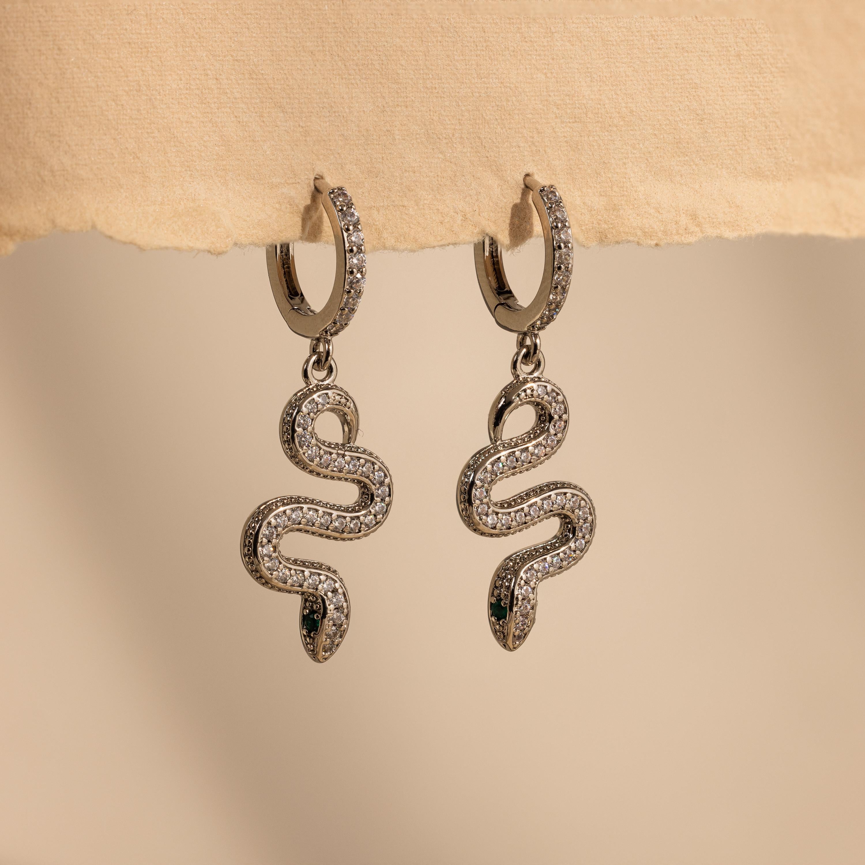 Pave Snake Charm Hoops, shaped like silver snakes with sparkling clear stones, dangle elegantly against a beige background.