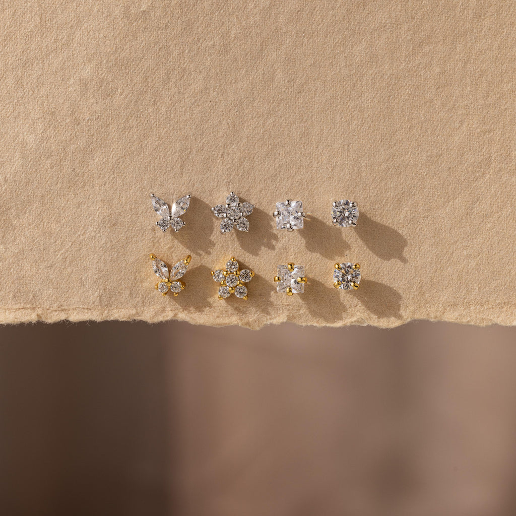 The Pave Assortment Flat Back Set features nine silver and gold pave diamond studs in butterfly, flower, and round designs, beautifully displayed on textured beige paper—an elegant earring set for any occasion.