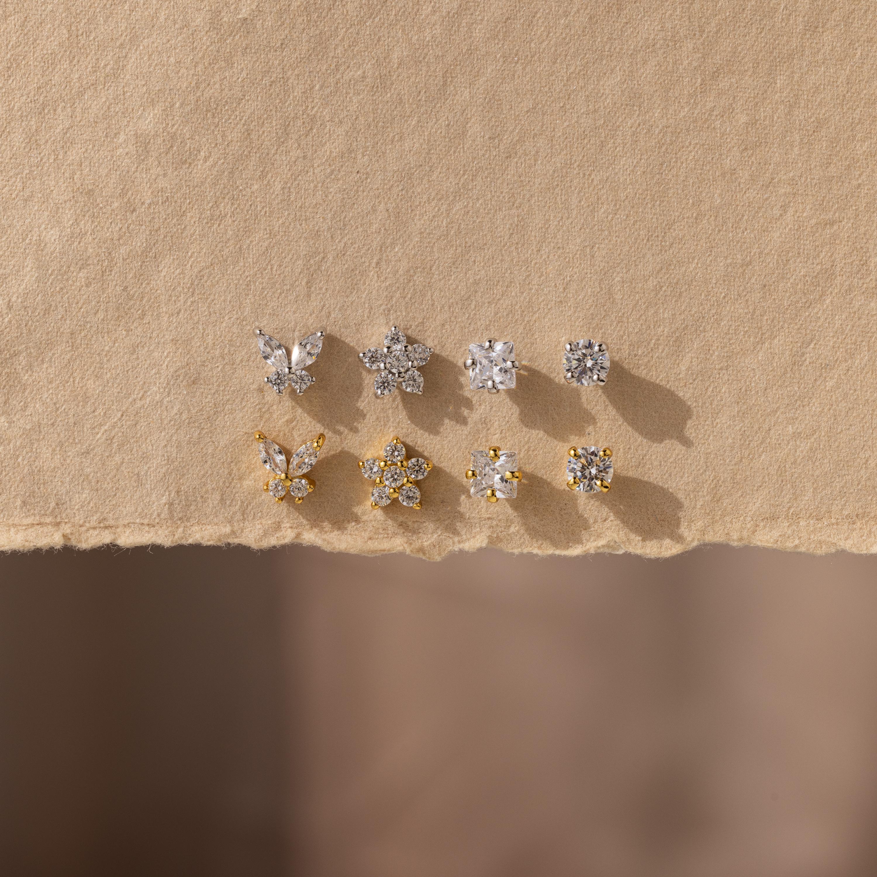 The Pave Assortment Flat Back Set features nine silver and gold pave diamond studs in butterfly, flower, and round designs, beautifully displayed on textured beige paper—an elegant earring set for any occasion.
