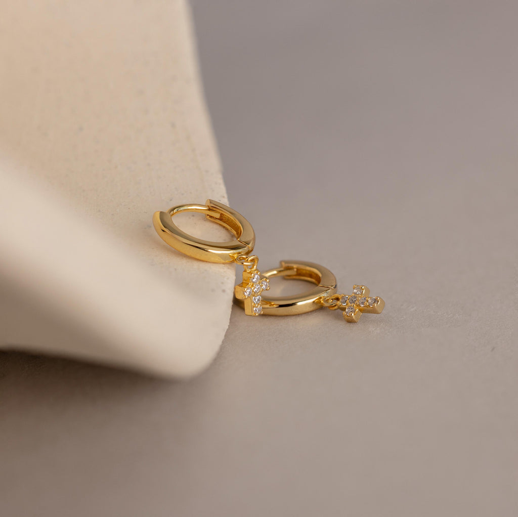 The Pave Cross Huggies, featuring a delicate pendant, are displayed on a soft beige surface.