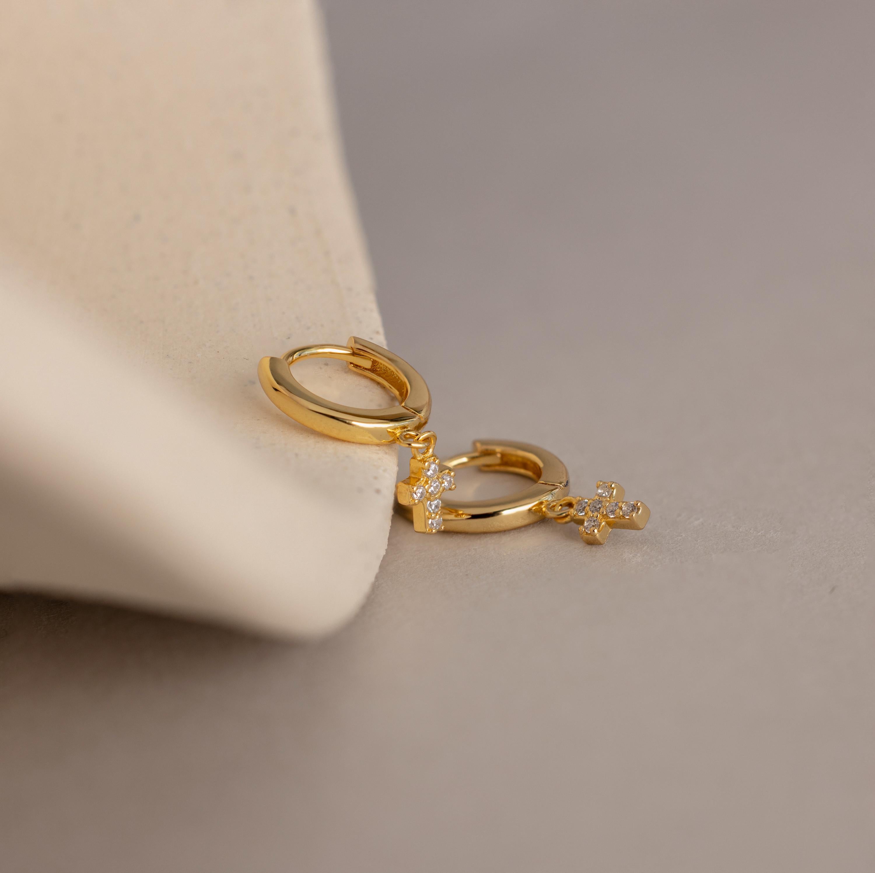 The Pave Cross Huggies, featuring a delicate pendant, are displayed on a soft beige surface.