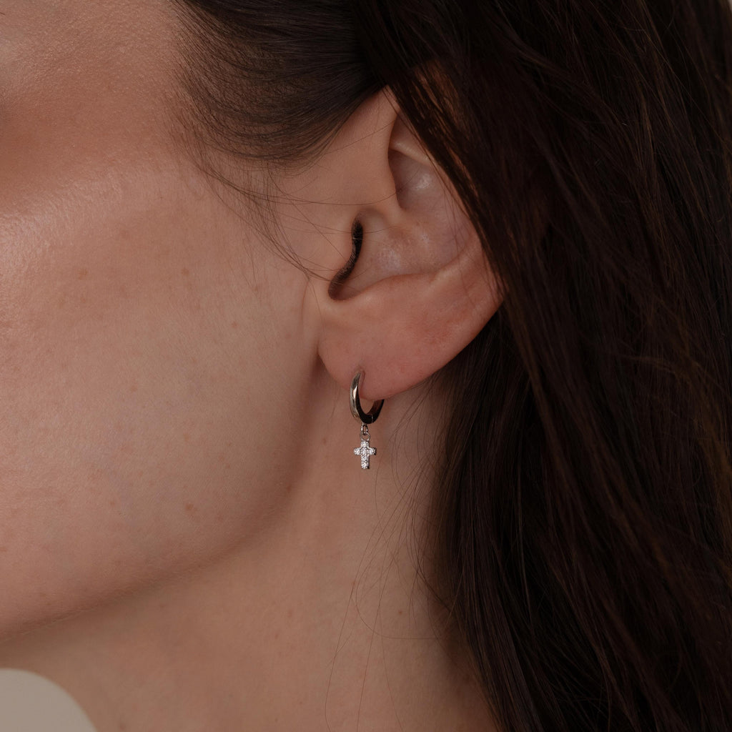 Close-up of a woman's ear wearing Pave Cross Huggies—small silver hoops with a dangling cross charm, perfect for a subtle yet elegant look.