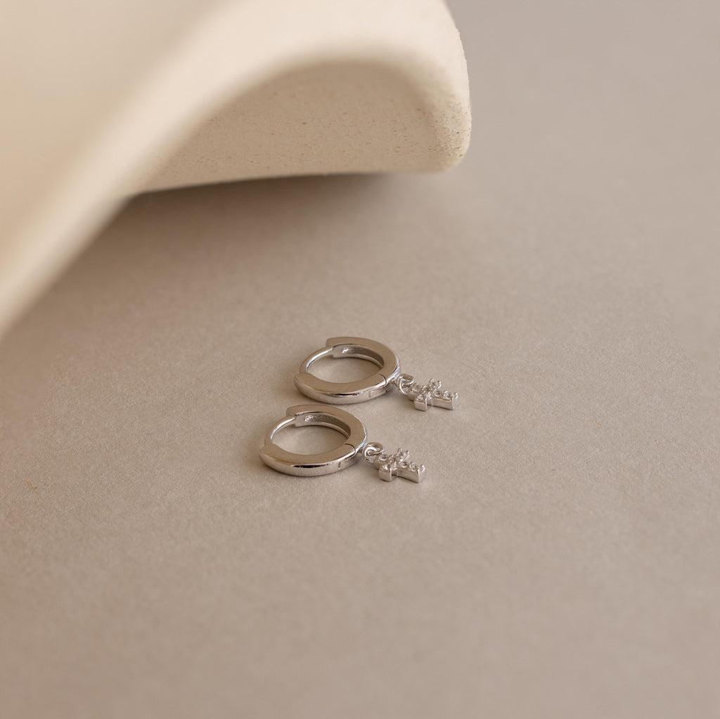 The Pave Cross Huggies—dainty hoops with small cross charms—add subtle sparkle, shown resting on a beige surface beside a curved, textured object.