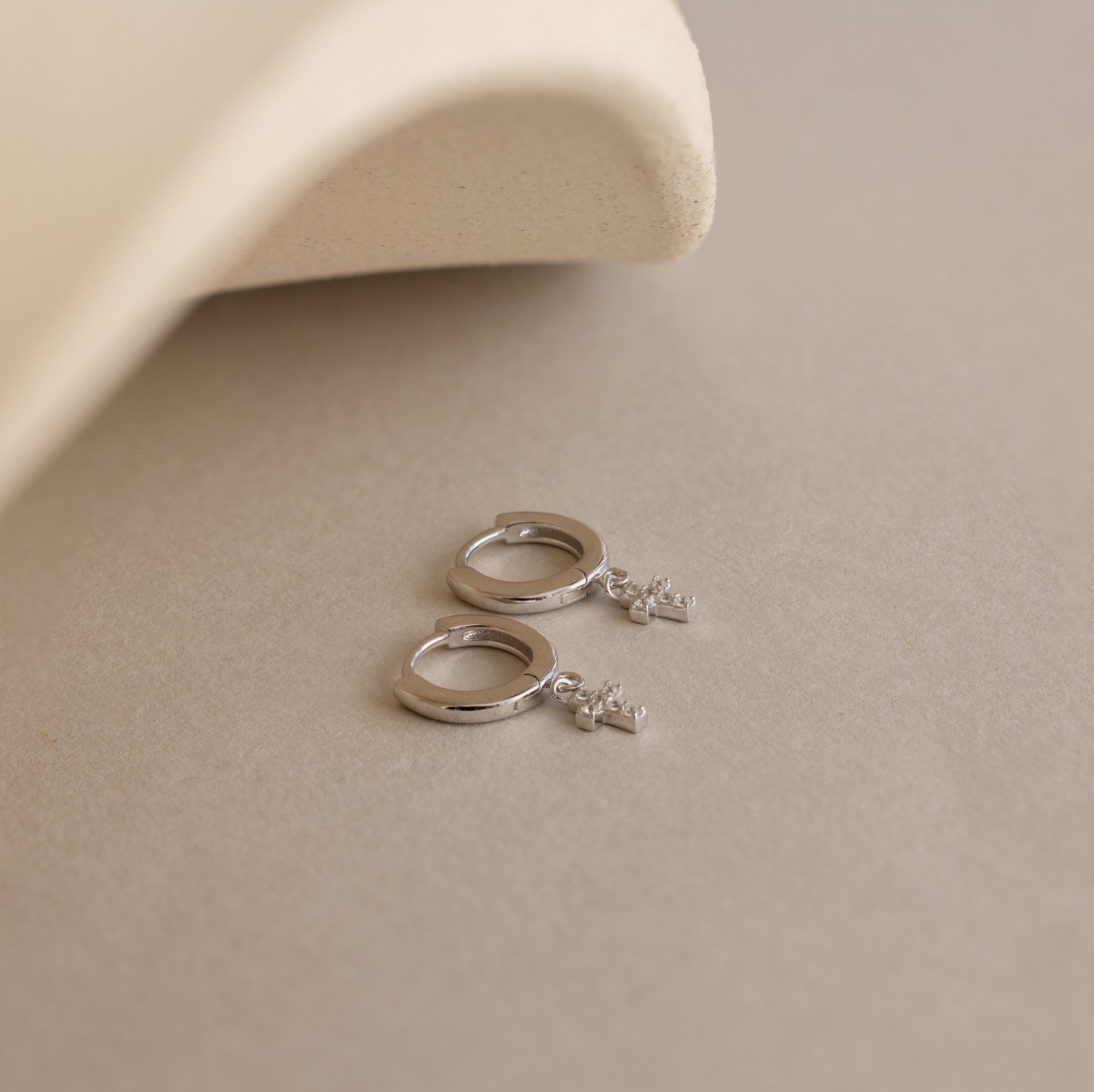 The Pave Cross Huggies—dainty hoops with small cross charms—add subtle sparkle, shown resting on a beige surface beside a curved, textured object.
