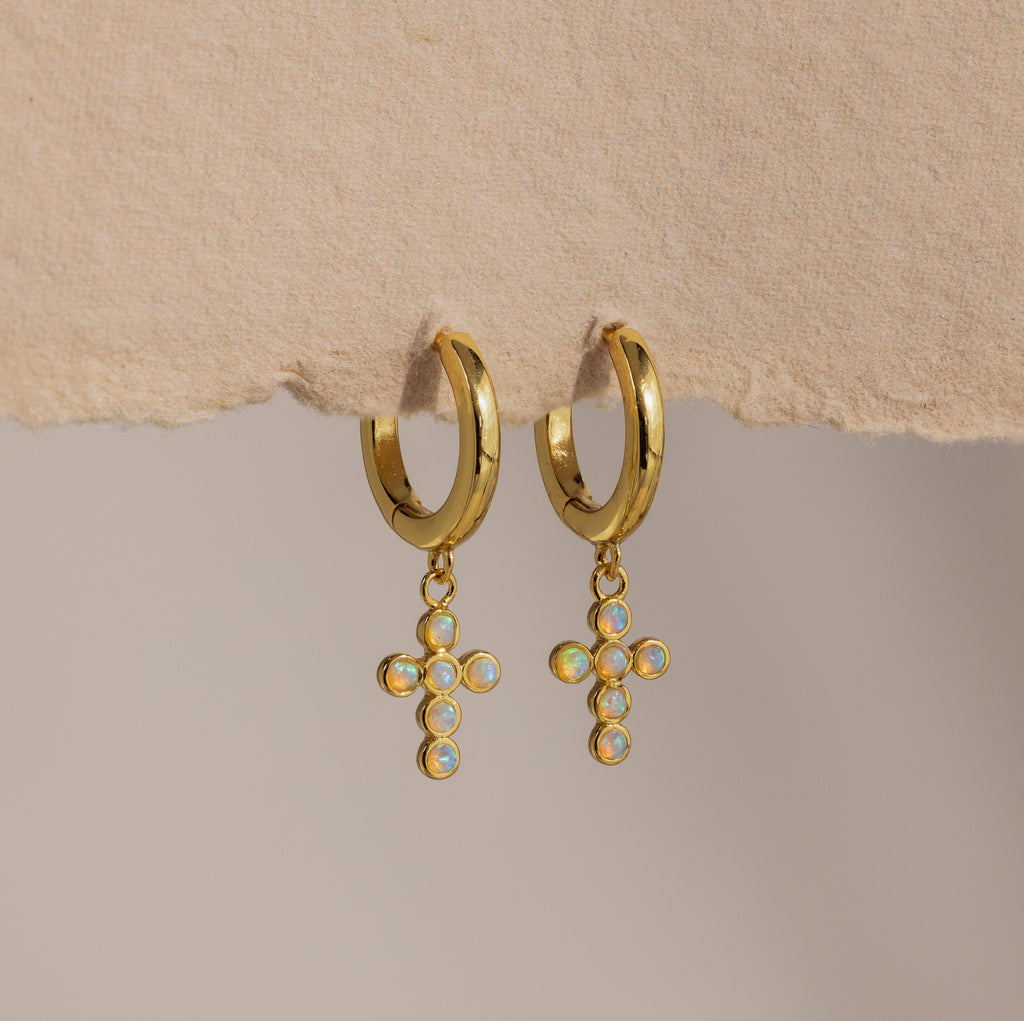 Opal Cross Huggies: gold huggie earrings with iridescent opal cross charms, shown on textured paper.