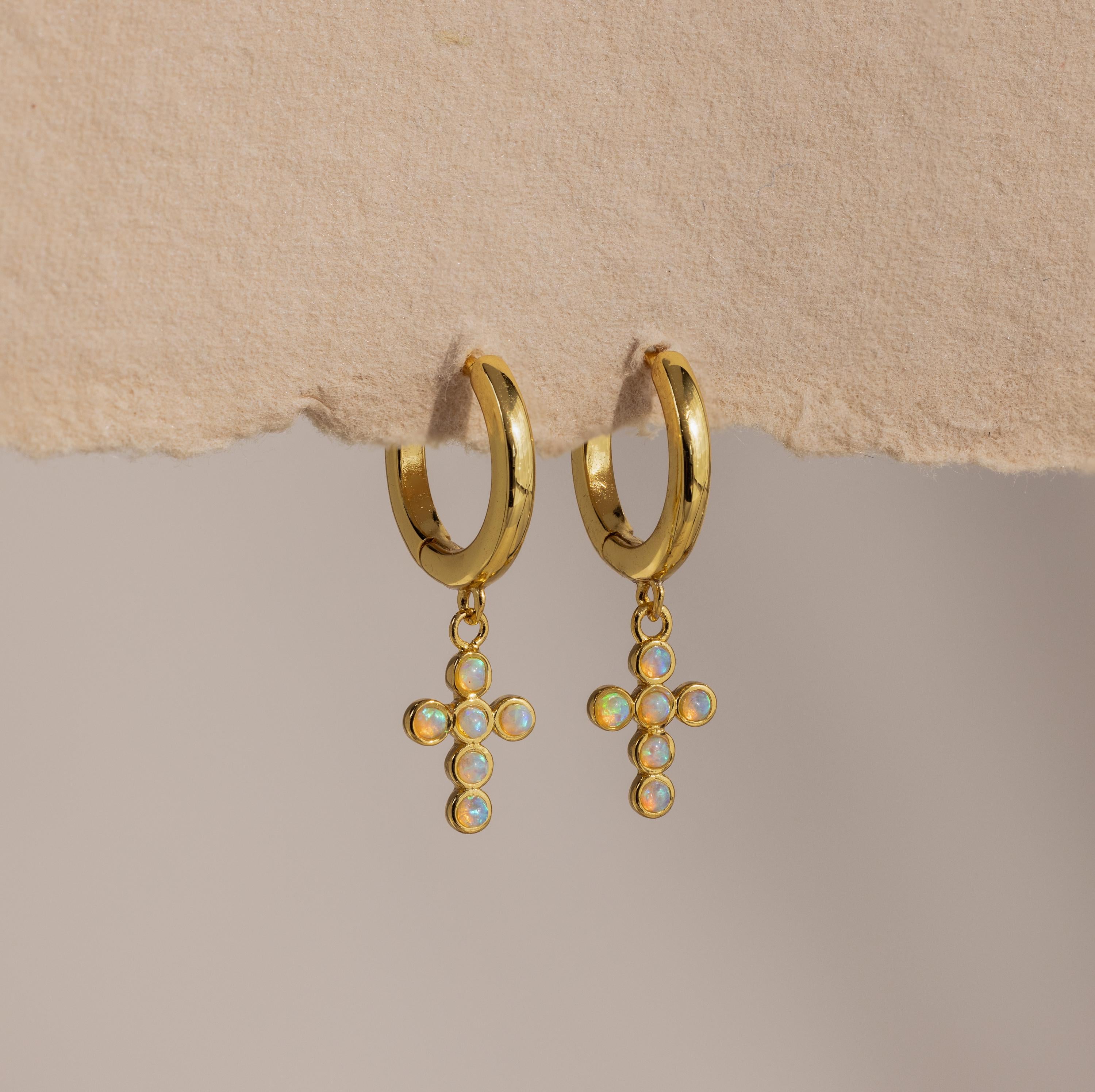 Opal Cross Huggies: gold huggie earrings with iridescent opal cross charms, shown on textured paper.