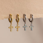 Opal Cross Huggies in gold and silver, featuring opal cross charms with gem-studded pendants, displayed on a beige surface.