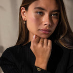 Woman with long brown hair wearing gold Opal Cross Huggies and a black top, resting her hand on her chin.