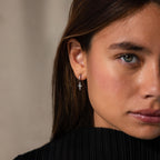 A woman models Opal Cross Huggies, showcasing their delicate opal cross charms. She stands against a neutral background with a serious expression.