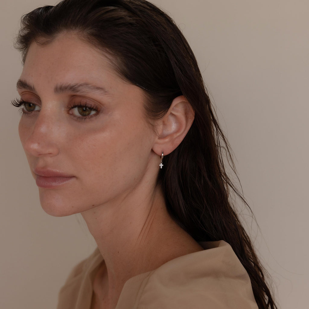 A woman with wet brown hair, dressed in a beige top, looks to the side while wearing delicate Pave Cross Huggies.
