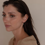A woman with wet brown hair, dressed in a beige top, looks to the side while wearing delicate Pave Cross Huggies.